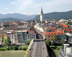 Eine Brücke führt über einen Fluss zu einer Stadt mit bunten Gebäuden und einem hohen Kirchturm vor der Kulisse der Berge.