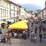 Eine belebte Fußgängerzone mit Geschäften und Cafés, in der die Menschen spazieren gehen und unter gelben Sonnenschirmen im Freien sitzen.