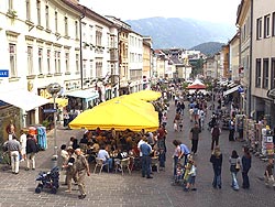 Eine belebte Fußgängerzone mit Geschäften und Cafés, in der die Menschen spazieren gehen und unter gelben Sonnenschirmen im Freien sitzen.