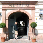 Eine Frau steht am Eingang eines Museums mit einem gewölbten Portal, flankiert von zwei Topfpflanzen und zwei an der Wand befestigten Lampen.