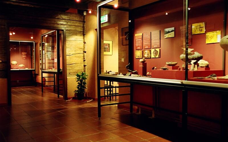 Innenraum des Museums mit Glasvitrinen, in denen Artefakte ausgestellt sind, und Informationstafeln an den Wänden, die mit warmem Licht beleuchtet werden.