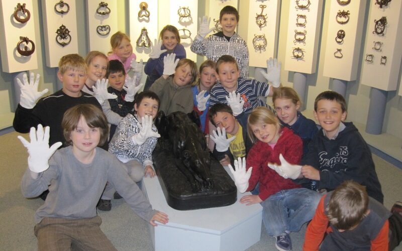 Eine Gruppe von Kindern mit weißen Handschuhen posiert mit einer schwarzen Statue in einem Museum, hinter der dekorative Gegenstände an den Wänden ausgestellt sind.