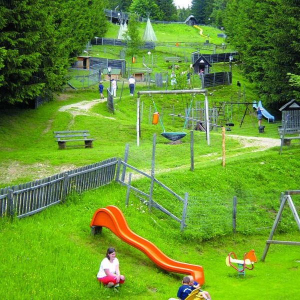 Kinder spielen in der Nähe einer roten Rutsche und einer Schaukel auf einem grasbewachsenen Spielplatz, der von Holzzäunen und Bäumen umgeben ist, während auf einem Hügel im Hintergrund weitere Spielgeräte zu sehen sind.