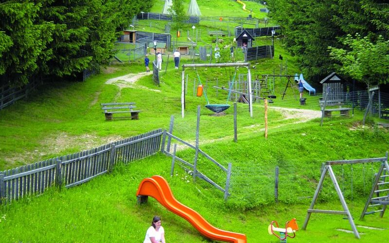 Kinder spielen in der Nähe einer roten Rutsche und einer Schaukel auf einem grasbewachsenen Spielplatz, der von Holzzäunen und Bäumen umgeben ist, während auf einem Hügel im Hintergrund weitere Spielgeräte zu sehen sind.