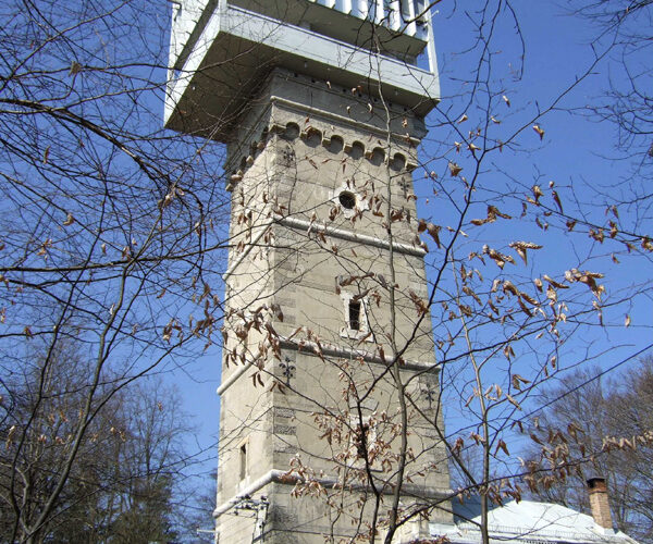 Ein hoher, rechteckiger Steinturm mit einer großen weißen Struktur auf der Spitze, umgeben von blattlosen Bäumen und einem geparkten Auto, unter einem klaren blauen Himmel.