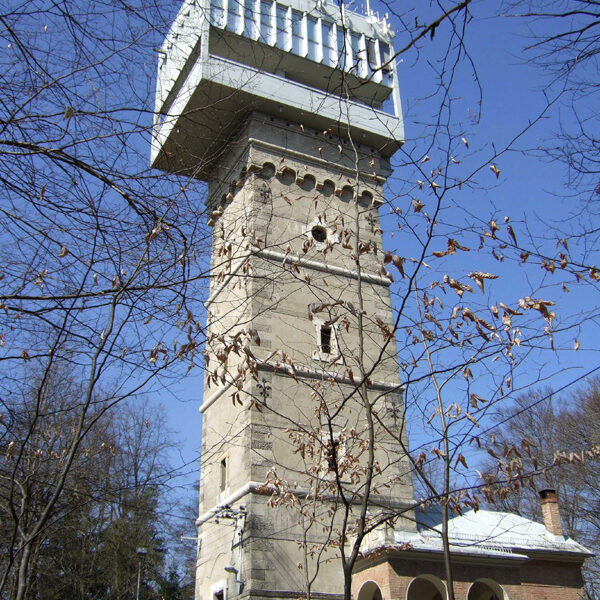 Ein hoher, rechteckiger Steinturm mit einer großen weißen Struktur auf der Spitze, umgeben von blattlosen Bäumen und einem geparkten Auto, unter einem klaren blauen Himmel.