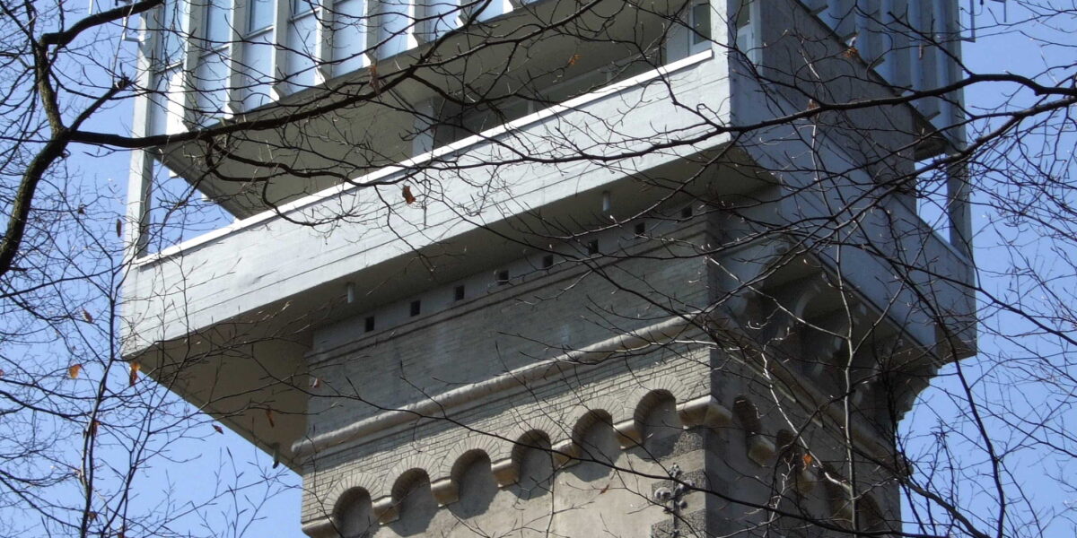 Ein hoher Steinturm mit einer modernen gläsernen Aussichtsplattform auf der Spitze, die man durch kahle Äste vor einem klaren blauen Himmel sieht.