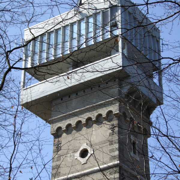 Ein hoher Steinturm mit einer modernen gläsernen Aussichtsplattform auf der Spitze, die man durch kahle Äste vor einem klaren blauen Himmel sieht.