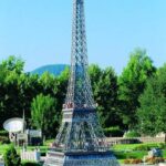 Das maßstabsgetreue Modell des Eiffelturms steht unter freiem Himmel, umgeben von Grün und anderen Miniaturgebäuden, unter einem klaren blauen Himmel.