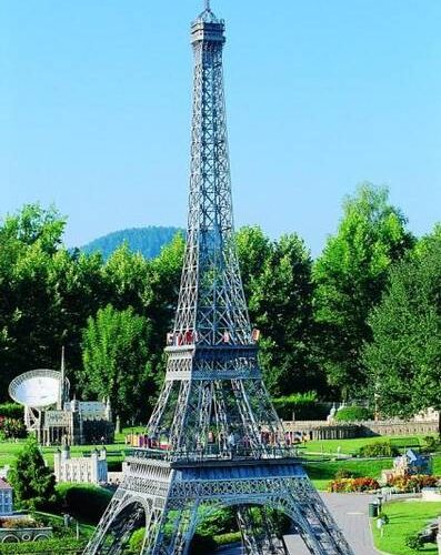 Das maßstabsgetreue Modell des Eiffelturms steht unter freiem Himmel, umgeben von Grün und anderen Miniaturgebäuden, unter einem klaren blauen Himmel.