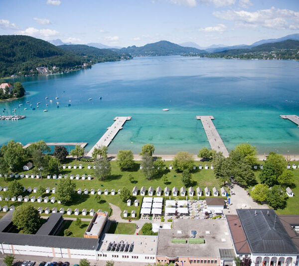 Luftaufnahme eines Seeufers mit klarem, blauem Wasser, mehreren Anlegestellen, grünen Parkanlagen, kleinen Gebäuden und umliegenden Hügeln unter einem teilweise bewölkten Himmel.