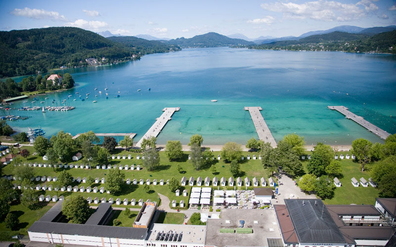 Luftaufnahme eines Seeufers mit klarem, blauem Wasser, mehreren Anlegestellen, grünen Parkanlagen, kleinen Gebäuden und umliegenden Hügeln unter einem teilweise bewölkten Himmel.