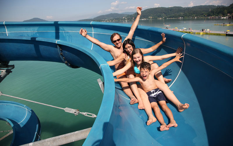 Vier Personen in Badeanzügen fahren gemeinsam eine blaue Wasserrutsche über ein großes Gewässer hinunter, mit Bergen und einem teilweise bewölkten Himmel im Hintergrund.