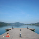 Zwei Personen sitzen auf einem hölzernen Steg an einem ruhigen See, umgeben von Bergen und unter einem strahlend blauen Himmel.