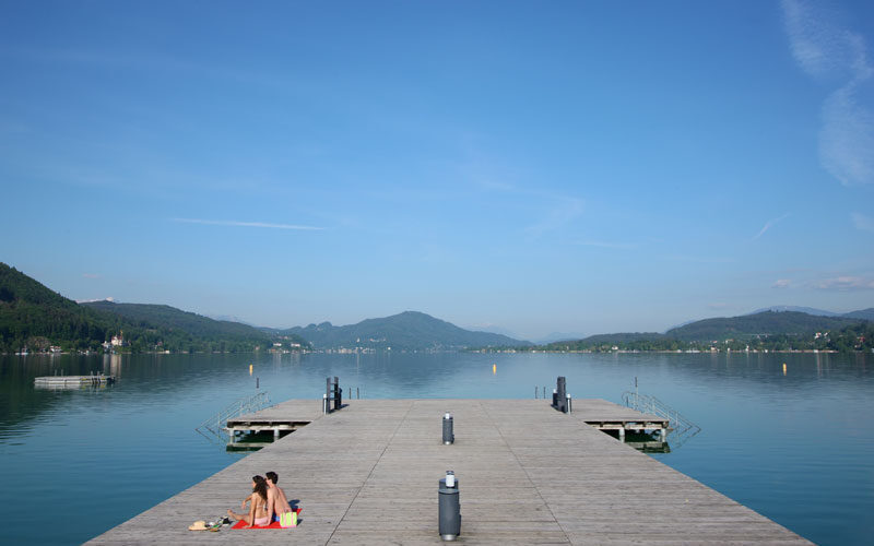 Zwei Personen sitzen auf einem hölzernen Steg an einem ruhigen See, umgeben von Bergen und unter einem strahlend blauen Himmel.