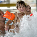 Drei Personen, darunter ein Kind mit orangefarbenen Schwimmflügeln, lächeln und spielen zusammen in einem Schwimmbad, während das Wasser um sie herum aufspritzt.