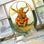 Drei Personen, zwei Erwachsene und ein Kind, rutschen lächelnd und mit erhobenen Armen eine Indoor-Wasserrutsche hinunter in ein Schwimmbecken.