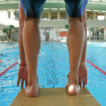Ein Schwimmer steht auf einem Startblock und bereitet sich auf den Sprung in ein Hallenbad vor, während im Hintergrund andere schwimmen.