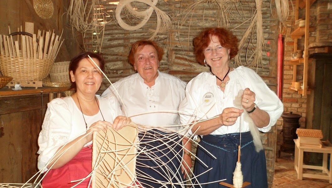 Drei Frauen in traditioneller Kleidung flechten Körbe aus hellem Schilf in einer rustikalen Werkstatt mit Körben und Flechtmaterial an den Wänden.