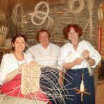 Drei Frauen in traditioneller Kleidung flechten Körbe aus hellem Schilf in einer rustikalen Werkstatt mit Körben und Flechtmaterial an den Wänden.