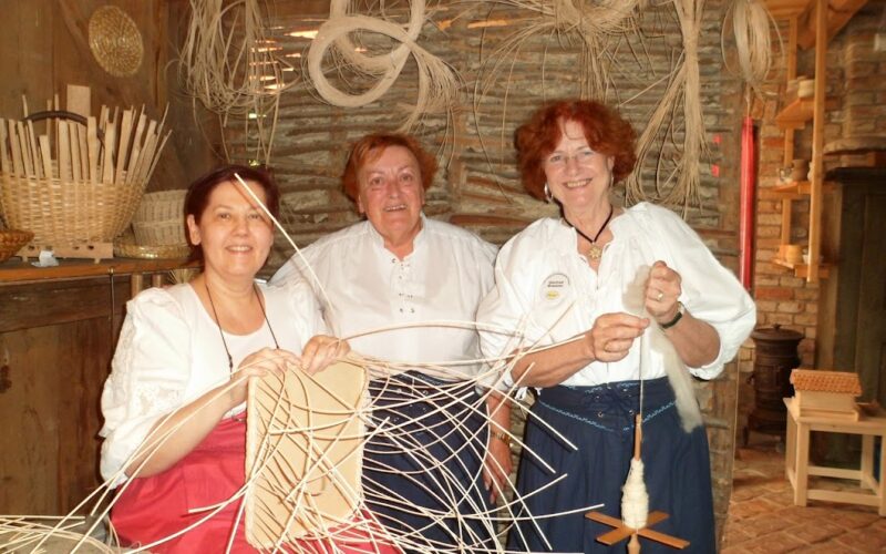 Drei Frauen in traditioneller Kleidung flechten Körbe aus hellem Schilf in einer rustikalen Werkstatt mit Körben und Flechtmaterial an den Wänden.