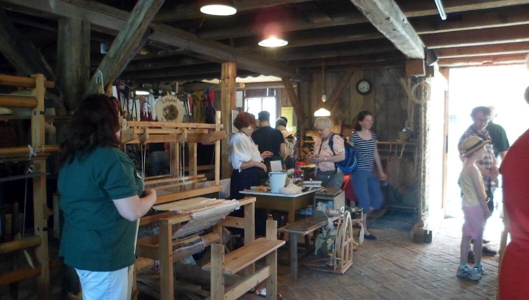 In einer rustikalen Holzwerkstatt mit Webstühlen, Spinnrädern und Kunsthandwerk, das auf Tischen und an den Wänden ausgestellt ist, stehen die Menschen und tauschen sich aus.