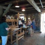 In einer rustikalen Holzwerkstatt mit Webstühlen, Spinnrädern und Kunsthandwerk, das auf Tischen und an den Wänden ausgestellt ist, stehen die Menschen und tauschen sich aus.