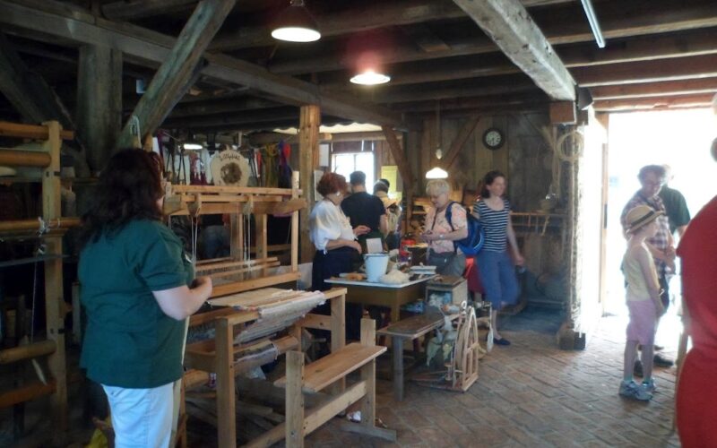 In einer rustikalen Holzwerkstatt mit Webstühlen, Spinnrädern und Kunsthandwerk, das auf Tischen und an den Wänden ausgestellt ist, stehen die Menschen und tauschen sich aus.