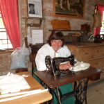 Eine Frau sitzt an einer alten Nähmaschine in einem rustikalen Raum mit Backsteinwänden, Holzmöbeln und Nähmaterial auf den Tischen.