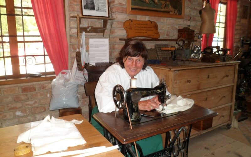 Eine Frau sitzt an einer alten Nähmaschine in einem rustikalen Raum mit Backsteinwänden, Holzmöbeln und Nähmaterial auf den Tischen.