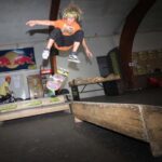 Ein Skateboarder in einem orangefarbenen Hemd vollführt einen Trick in der Luft auf einer Holzrampe in einem Indoor-Skatepark.