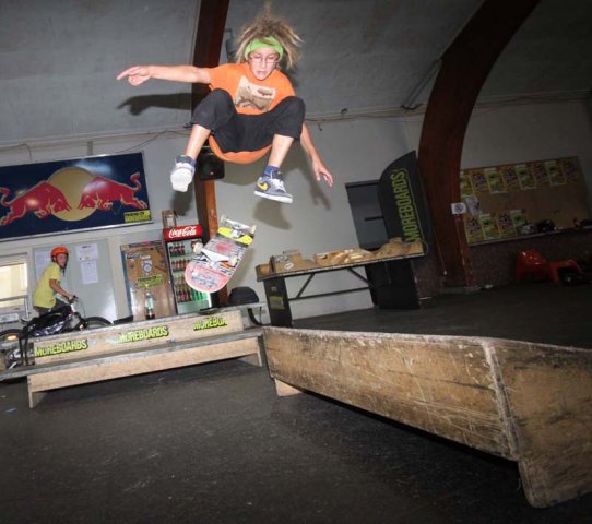 Ein Skateboarder in einem orangefarbenen Hemd vollführt einen Trick in der Luft auf einer Holzrampe in einem Indoor-Skatepark.