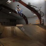 Eine Person fährt mit einem BMX-Rad von einer Rampe in einem Indoor-Skatepark und wirft dabei einen Schatten auf die gewölbte Wand hinter ihr.
