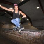 Eine Person in einem schwarzen Tank-Top, blauen Jeans und einer roten Kappe fährt mit Rollschuhen auf einer Rampe in einem Skatepark, wobei sie die Arme zum Ausgleich ausstreckt.