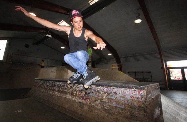 Eine Person in einem schwarzen Tank-Top, blauen Jeans und einer roten Kappe fährt mit Rollschuhen auf einer Rampe in einem Skatepark, wobei sie die Arme zum Ausgleich ausstreckt.