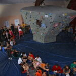 Boulder-Wettbewerb in der Halle mit Kletterern an einer Kletterwand und einer Menge von Zuschauern, die auf blauen Matten sitzen und stehen.