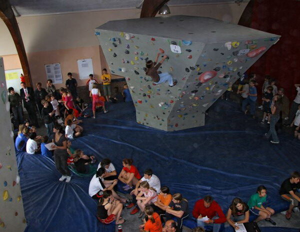 Boulder-Wettbewerb in der Halle mit Kletterern an einer Kletterwand und einer Menge von Zuschauern, die auf blauen Matten sitzen und stehen.