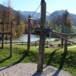 Ein Klettergerüst für einen Spielplatz im Freien mit Holzpfosten und roten Seilen, auf Gras und Kies, mit Bergen und Bäumen im Hintergrund.