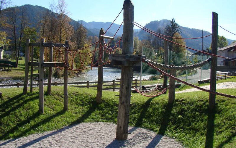 Ein Klettergerüst für einen Spielplatz im Freien mit Holzpfosten und roten Seilen, auf Gras und Kies, mit Bergen und Bäumen im Hintergrund.
