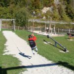 Ein Kind in einer rot-schwarzen Jacke fährt mit einer Seilrutsche auf einem Spielplatz mit Gras, Kieswegen und Spielgeräten.