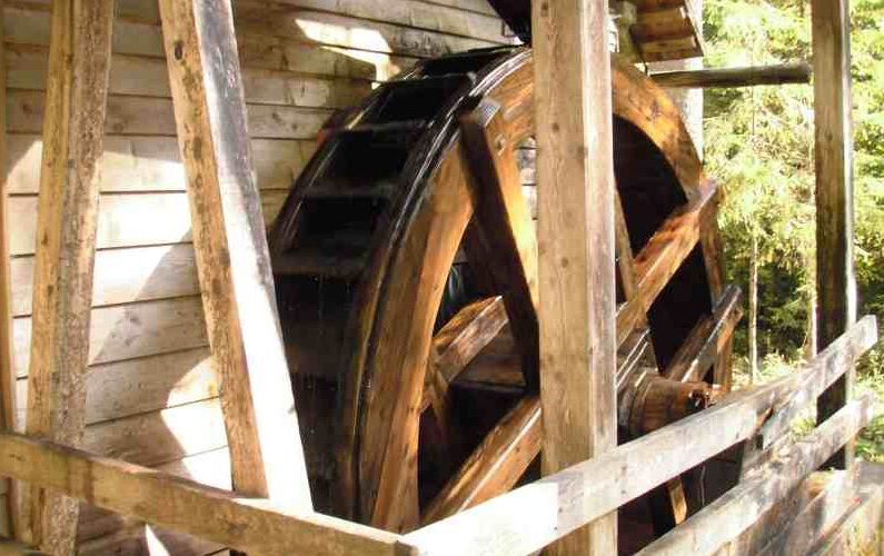 Ein großes hölzernes Wasserrad ist an der Seite eines rustikalen Holzgebäudes angebracht, das von Holzbalken und Geländern umgeben ist.