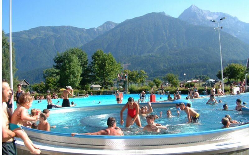 Menschen verschiedenen Alters schwimmen und spielen in einem Freibad, umgeben von Grün und Bergen unter einem klaren Himmel.