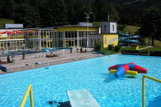 Freibad mit einem bunten aufblasbaren Spielzeug, einem gelben Sprungbrett und einem modernen Gebäude mit blauen Wasserrutschen im Hintergrund, umgeben von Bäumen und Grünanlagen.