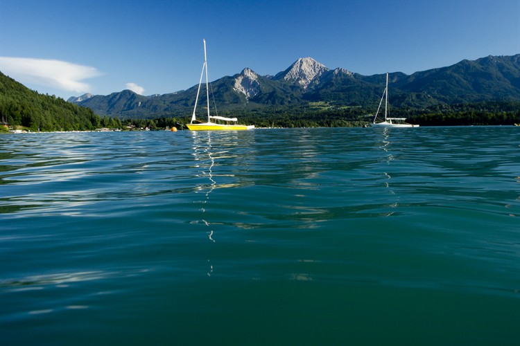 Zwei Segelboote treiben auf einem ruhigen See mit klarem Wasser, umgeben von grünen Hügeln und Bergen unter einem strahlend blauen Himmel.