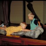 Zwei kleine Kinder interagieren mit einem großen Dinosaurierknochen, der auf einer hölzernen Plattform in einer Museumsausstellung ausgestellt ist.