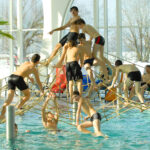 Eine Gruppe von Jungen in Badekleidung klettert und spielt auf einer Seilstruktur über einem Becken in einem Indoor-Badezentrum mit großen Fenstern und einer Palme.