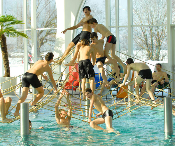 Eine Gruppe von Jungen in Badekleidung klettert und spielt auf einer Seilstruktur über einem Becken in einem Indoor-Badezentrum mit großen Fenstern und einer Palme.