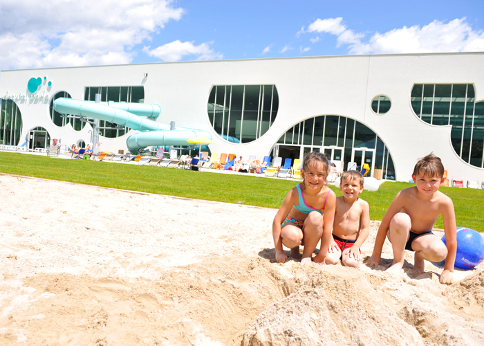 Drei Kinder in Badeanzügen knien an einem sonnigen Tag auf sandigem Boden vor einem modernen Gebäude mit großen Fenstern und Wasserrutschen im Freien.