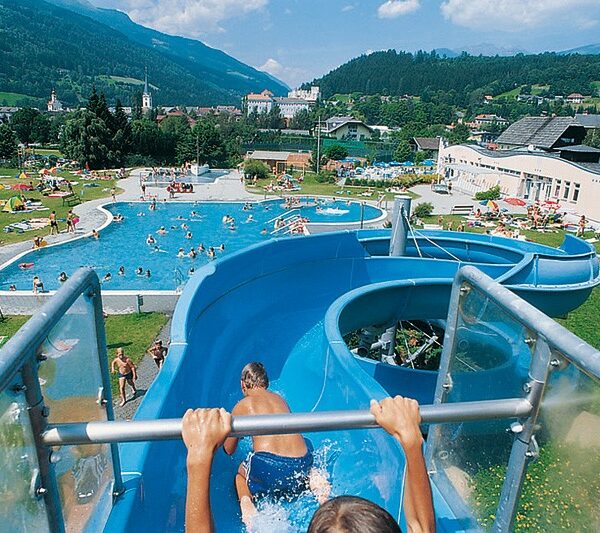 Zwei Kinder rutschen eine blaue Wasserrutsche hinunter in ein Freibad, umgeben von Menschen, Grasflächen und Bergen im Hintergrund.