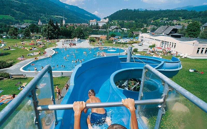 Zwei Kinder rutschen eine blaue Wasserrutsche hinunter in ein Freibad, umgeben von Menschen, Grasflächen und Bergen im Hintergrund.